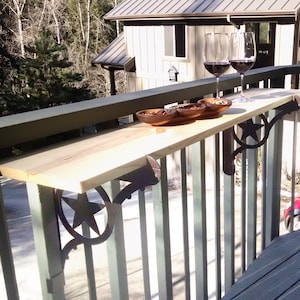 May include: A wooden bar table with a star-shaped metal bracket attached to a railing. The table is made of light wood and has a natural finish. Two glasses of red wine are on the table, along with a bowl of nuts.