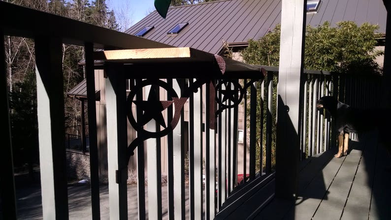 May include: A black metal railing with a wooden top and star-shaped accents. The railing is attached to a wooden deck and a house in the background.