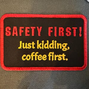 Könnte beinhalten: Ein rechteckiger schwarzer Aufnäher mit rotem Rand und dem Text "SAFETY FIRST!" in Rot, gefolgt von "Just kidding, coffee first." in Gelb. Der Aufnäher ist wahrscheinlich ein Accessoire.