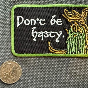 May include: A black embroidered patch with a green border and the words "Don't be hasty" in white script. The patch features a stylized illustration in yellow and green. A quarter coin is next to the patch.