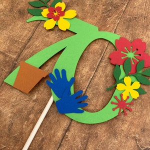 Può includere: Un cake topper verde a forma di numero sette con un vaso di fiori marrone, mani blu e fiori di carta colorati. I fiori sono rossi, gialli e verdi. Il cake topper è attaccato a un bastoncino bianco.
