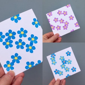 Peut inclure: Trois cartes blanches avec des motifs floraux. L'une présente des fleurs bleues à centres jaunes, une autre des fleurs violettes à centres jaunes, et la dernière des fleurs bleu clair à centres jaunes. Les cartes sont tenues par des mains.