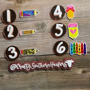 May include: A set of six brown and white circle-shaped number stickers with a white outline. Each number is paired with a different school-themed image, including a rainbow pencil, a leopard print pencil, a leopard print pencil, a pink bow pencil, a pink heart pencil, and a set of rainbow crayons. The stickers are on a wooden background. The text "@CraftySouthernHerfers" is on a pink glitter background.
