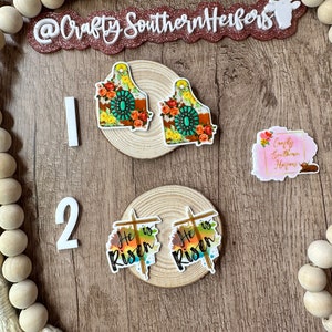 May include: Two sets of earrings on a wooden background. The first set features a floral design with turquoise accents. The second set features a watercolor design with the words "He is Risen" and a cross.