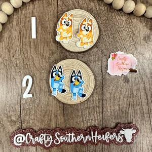 May include: Two sets of character cutouts on wooden circles. The first set features two orange and white cartoon dogs with blue eyes. The second set features two blue and white cartoon dogs with red eyes. The text "Crafty Southern Heifers" is on a pink glitter background.