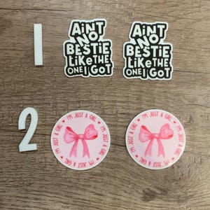 May include: Two sets of stickers. One set says "Ain't no bestie like the one I got" in black text on a white background. The other set is a pink circle with a pink bow and the text "I'm just a girl" in black text.