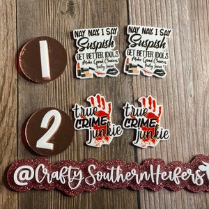 May include: Two sets of round, white stickers with black text. The first set says "Nay Nay, I Say Suspish Get Better Idols Make Good Choices" with a lipstick and a bloody knife graphic. The second set says "True Crime Junkie" with a bloody handprint graphic. Two round, copper-colored stickers with white numbers "1" and "2". A pink glitter banner with the text "@Crafty Southern Herfers" and a white cow silhouette.