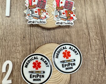 May include: Two white round stickers with black borders and red medical alert symbols. The text "MEDICAL ALERT" is printed in black around the symbol. The text "EPIPEN INSIDE" is printed in black below the symbol. The stickers are on a wooden surface.