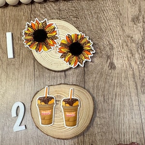 May include: Two wood slices with decorative stickers. The first slice has two sunflower stickers with a leopard print design. The second slice has two stickers of iced coffee cups with the text "Dunkin' Donuts".
