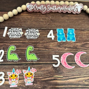 May include: A set of colorful stickers featuring a variety of designs, including dinosaurs, gummy bears, unicorns, and moons. The stickers are arranged on a wooden surface with a string of wooden beads.