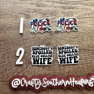 May include: Two white numbers, "1" and "2", are displayed on a wooden surface.  Two white and blue police car themed epoxy resin ornaments with the text "Police Life" are displayed above the numbers.  Two white epoxy resin ornaments with the text "Somebody's Spoiled Blue Collar Wife" are displayed below the numbers.  A red glitter epoxy resin ornament with the text "@Crafty Southern Henfers" is displayed below the ornaments.