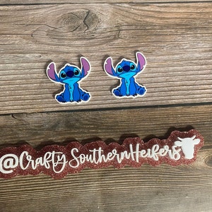 May include: Two blue Stitch character stickers with purple ears and white outlines. The stickers are on a wooden background with a pink glitter sign that says "@CraftySouthernHeifers" with a white cow silhouette.
