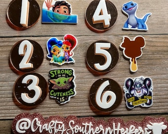 May include: Six round brown and white circles with numbers 1 through 6. Each circle has a different character from a Disney movie. The characters are: Luca,  Shimmer and Shine, Baby Yoda,  Olaf,  Elsa, and Ursula.  The circles are on a wooden background.  The text "@Crafty Southern Herfers" is below the circles.