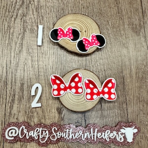 May include: Two sets of red and white polka dot Minnie Mouse ears with black ears. The ears are on a wooden background with the numbers 1 and 2 and the text "@Crafty Southern Herfers".
