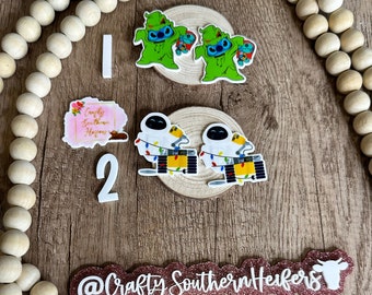 May include: A set of four Christmas-themed resin ornaments. Two ornaments feature a green Stitch character wearing a Santa hat. The other two ornaments feature a white robot character with Christmas lights. The ornaments are on a wooden surface with a pink and white sign that says "Crafty Southern Heifers".