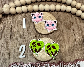 May include: Two pairs of novelty earrings. One pair features a pink cartoon character with blue eyes, and the other pair depicts a green apple with a dripping, dark brown design. The earrings are displayed on small wooden slices.