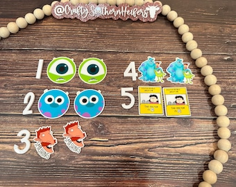 May include: A set of five white numbers, 1 through 5, with various cartoon character stickers. The stickers include two green monsters with one eye each, two blue monsters with two eyes each, and two red-haired characters with the word "smashing" written below them. The stickers are on a wooden background with a string of wooden beads.