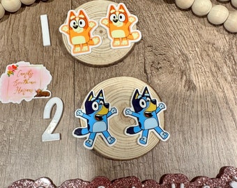 May include: Two sets of cartoon character stickers, one orange and one blue, are displayed on wooden slices. The stickers are arranged with the numbers 1 and 2, and a decorative sign with the text "Crafty Southern Heifers". Wooden beads are also present.