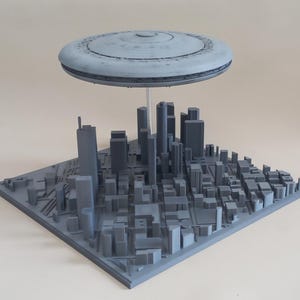May include: A gray 3D-printed model of a city with a hovering UFO. The city model features numerous rectangular buildings and a square base. The UFO is a disc shape with a detailed top. The model is a unique decorative item.
