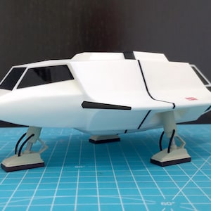 May include: A white model spaceship with black detailing and a red logo. The spaceship has a futuristic design with a cockpit and landing gear. The model is set against a blue grid background.