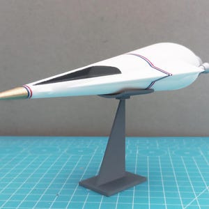 Könnte beinhalten: Weißes Modell eines futuristischen Flugzeugs mit goldfarbener Nase und schwarzem Fenster. Das Flugzeug hat rote und blaue Streifen an den Seiten und ist auf einem grauen Ständer montiert. Das Modell wird vor einem blauen Gitterhintergrund präsentiert.