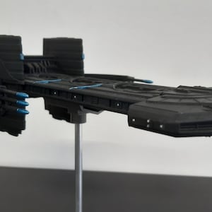May include: A black spaceship model with blue accents. The model is on a stand and has a long, narrow body with a rounded front and a pointed back.