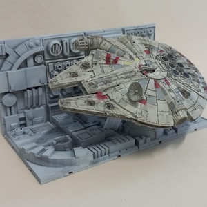 May include: A detailed model of the Millennium Falcon starship, with a weathered, grey and white finish, and red accents. The ship is positioned within a grey, industrial-style diorama, showcasing its iconic design. The diorama has detailed panels and structures.