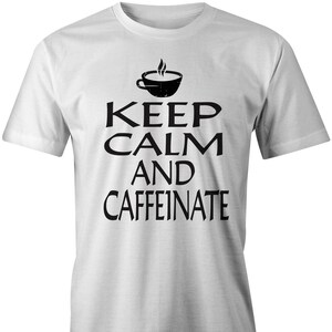 Op de afbeelding: Wit T-shirt met zwarte tekst die "Keep Calm and Caffeinate" luidt met een gestileerde koffiekopgrafiek boven de tekst.