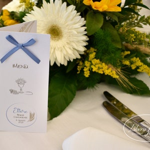May include: A white menu card with a blue ribbon tied around it. The card has a drawing of a chalice, bread, and grapes. The text on the card reads "MENU" and "Ettore Prima Comunione 6 Maggio 2023".