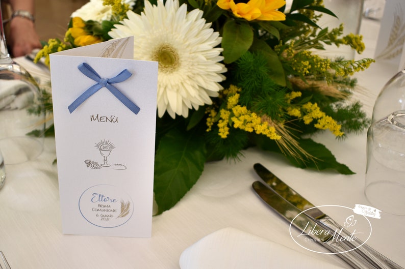 May include: A white menu card with a blue ribbon tied around it. The card has a drawing of a chalice, bread, and grapes. The text on the card reads "MENU" and "Ettore Prima Comunione 6 Maggio 2023".