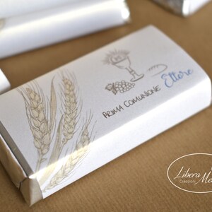 May include: White chocolate bar wrapped in silver foil with a white label. The label has a wheat design, a drawing of a chalice, bread, and grapes, and the text "Prima Comunione Ettore". The bottom of the label has the text "Libera Mente Creazioni".