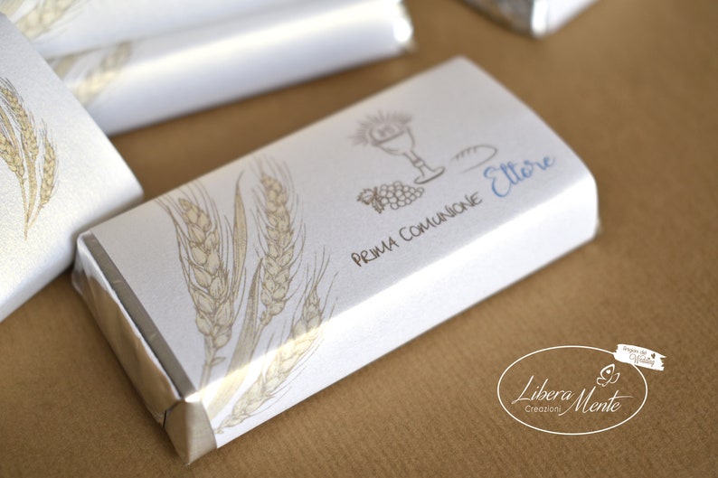 May include: White chocolate bar wrapped in silver foil with a white label. The label has a wheat design, a drawing of a chalice, bread, and grapes, and the text "Prima Comunione Ettore". The bottom of the label has the text "Libera Mente Creazioni".