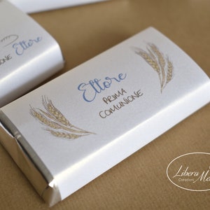 May include: A white chocolate bar wrapper with a silver foil border. The wrapper has a wheat design and the text "Ettore Prima Comunione".