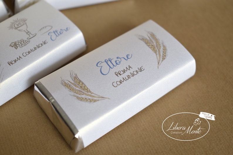 May include: A white chocolate bar wrapper with a silver foil border. The wrapper has a wheat design and the text "Ettore Prima Comunione".
