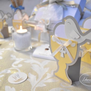 May include: A table setting for a special occasion with a white tablecloth and gold accents. There are several small white doves with yellow chalices and white ribbons. The table also has a small white candle in a clear glass holder. The text "Libera Mente Creazioni" is visible on the table.