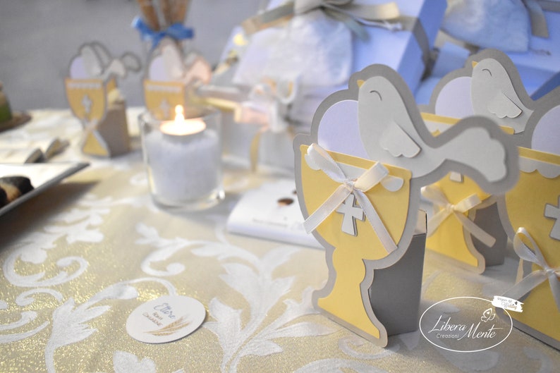 May include: A table setting for a special occasion with a white tablecloth and gold accents. There are several small white doves with yellow chalices and white ribbons. The table also has a small white candle in a clear glass holder. The text "Libera Mente Creazioni" is visible on the table.