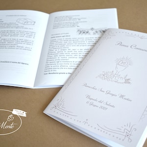 May include: A white booklet with a silver border and a silver illustration of a chalice with grapes and wheat. The text on the cover reads "Prima Comunione" and "Parrocchia San Giorgio Martire" with the date "6 Giugno 2021".