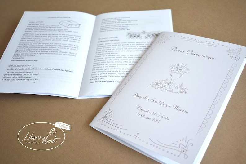 May include: A white booklet with a silver border and a silver illustration of a chalice with grapes and wheat. The text on the cover reads "Prima Comunione" and "Parrocchia San Giorgio Martire" with the date "6 Giugno 2021".