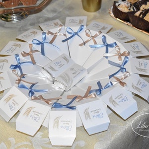May include: A white plate with a fan of white favor boxes tied with blue and brown ribbons. The boxes have a wheat design and the text "Grazie per la vostra presenza" in blue lettering.