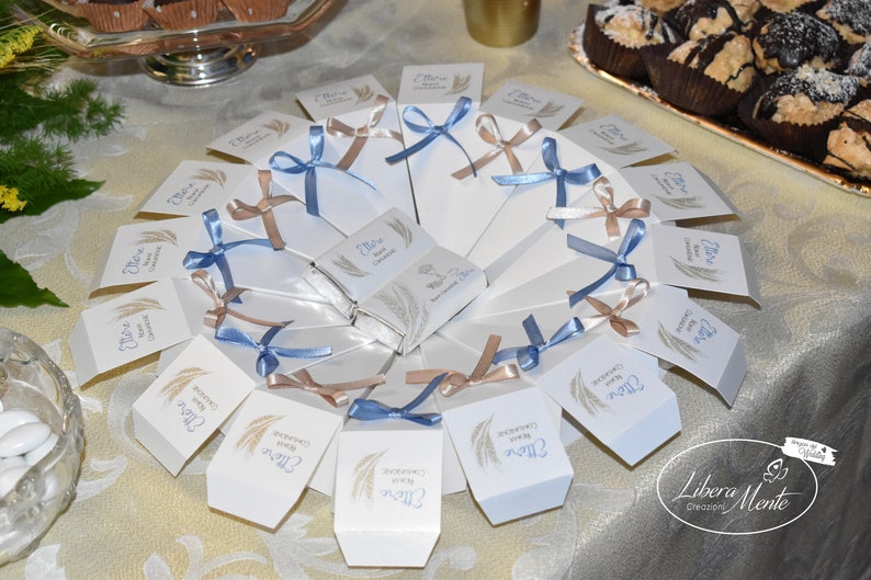 May include: A white plate with a fan of white favor boxes tied with blue and brown ribbons. The boxes have a wheat design and the text "Grazie per la vostra presenza" in blue lettering.