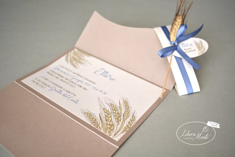May include: A beige invitation card with a wheat design and the text "Lettore" in blue. The card is folded in half and has a blue ribbon tied around it. The ribbon is attached to a small tag with the text "Lettore" in blue.