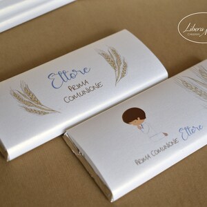 May include: Two white chocolate bar wrappers with the name "Ettore" and the text "Prima Comunione" printed on them. One wrapper features a drawing of a boy kneeling in prayer, while the other has a wheat stalk design.