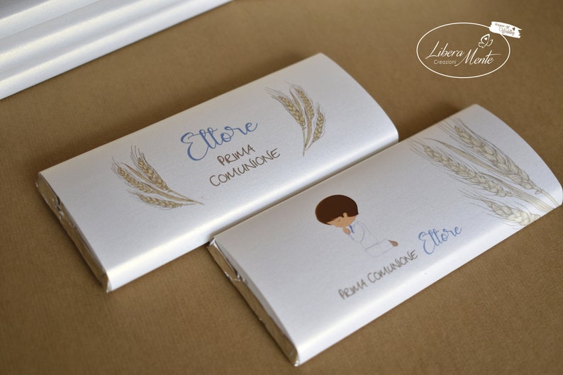 May include: Two white chocolate bar wrappers with the name "Ettore" and the text "Prima Comunione" printed on them. One wrapper features a drawing of a boy kneeling in prayer, while the other has a wheat stalk design.