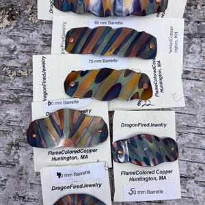 May include: A collection of flame-colored copper barrettes in various sizes, ranging from 40 mm to 80 mm. Each barrette features unique patterns of blues, greens, and golds, with copper rivets. The text "DragonFired Jewelry" is visible.