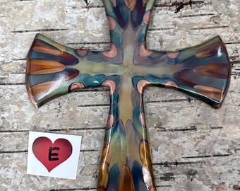 Può includere: Un oggetto decorativo a forma di croce con una superficie liscia e lucida caratterizzata da un motivo ripetitivo di petali allungati nei toni del verde acqua, rame e viola scuro. È visibile un piccolo adesivo quadrato con un cuore rosso e la lettera E.