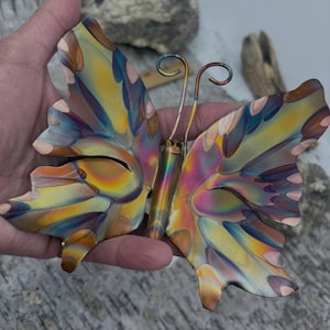 May include: A metal butterfly ornament featuring an iridescent finish with blended hues of yellow, blue, purple, and pink. The wings have a layered, scalloped edge design, and the body includes two thin, curved wire antennae extending from the top.