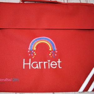 May include: Red book bag with a white rainbow and the name Harriet embroidered on it. The bag has white stripes on the side.