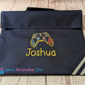 May include: A navy blue book bag with a white stripe on the side. The bag has a yellow embroidered video game controller and the name "Joshua" on the front. The text "Karen's Personalised Gifts" is on the bottom of the bag.