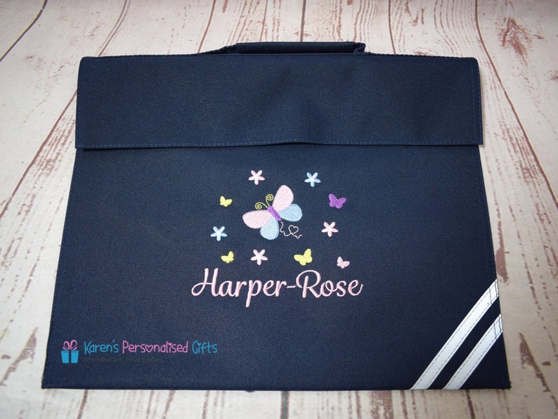 Puede incluir: Una mochila azul marino con rayas blancas a los lados. La mochila tiene un dise&ntilde;o de mariposa con detalles en rosa, amarillo y morado. El nombre "Harper-Rose" est&aacute; bordado en la mochila con hilo rosa.