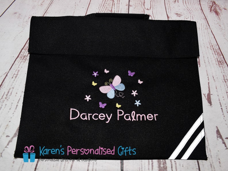 Puede incluir: Mochila negra para libros con rayas blancas en el lateral. La mochila tiene un dise&ntilde;o de mariposa con detalles en rosa, morado y amarillo. El nombre "Darcey Palmer" est&aacute; bordado en la mochila.